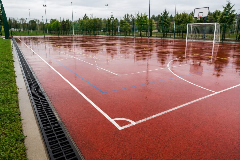 Suprafață sportivă multisport după ploaie, cu apă direcționată către o rigolă perimetrală de drenaj prin intermediul pantelor terenului